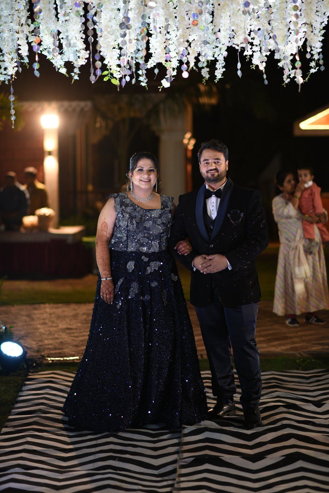 A photograph from the sangeet: the bride and groom in evening formal wear, caught mid-frame under a hanging white floral canopy on the night's chevron walkway, one frame out of the terabyte-plus of raw footage the rest of this piece is about wrangling.